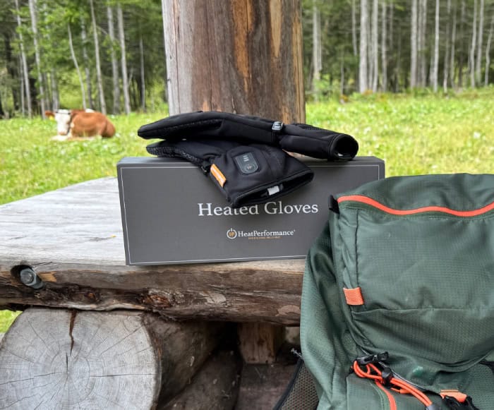 Beheizte Handschuhe von HeatPerformance® auf einer Holzbank während einer Wanderung, mit einem Rucksack und einer Bergwiese im Hintergrund.